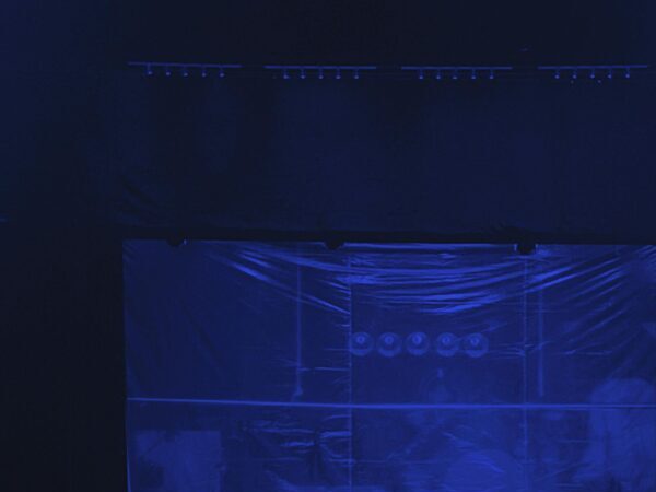 A dark stage with four empty chairs under low blue lighting, creating a moody atmosphere.